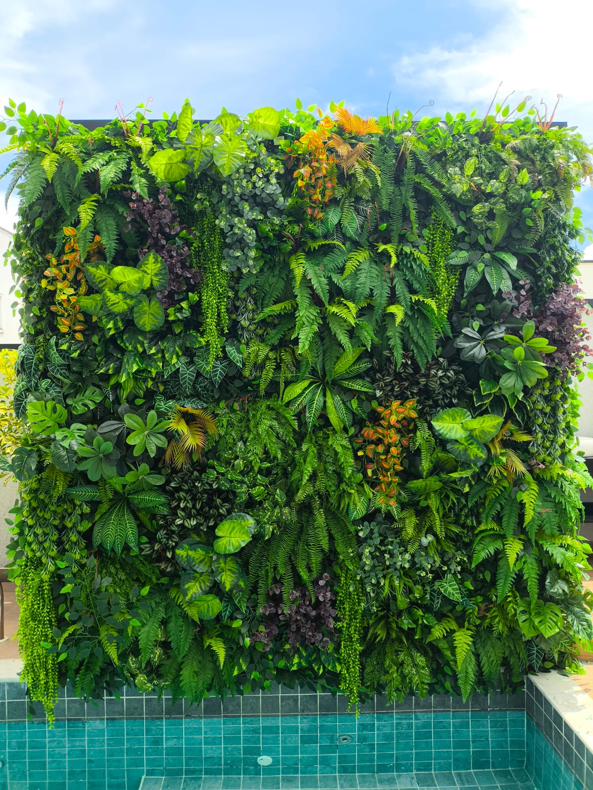 Parede de jardim vertical artificial densa e exuberante instalada em fachada residencial moderna, cobrindo toda a extensão vertical com folhagens variadas em tons de verde vivo — samambaias, pothos, monstera e trepadeiras — ao lado de piscina com azulejos turquesa, céu azul ao fundo, iluminação natural de dia ensolarado, fotografia realista de alto padrão, ângulo frontal